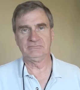 A middle-aged electrician with short gray hair wears a light blue collared shirt and looks directly at the camera with a neutral expression against a plain background.