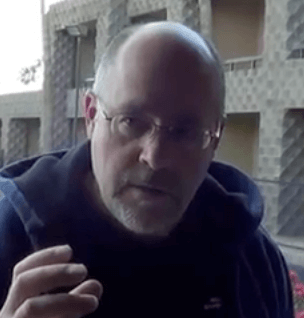 A bearded, middle-aged man wearing glasses looks at the camera, standing in front of a building with a geometric pattern that highlights the professionalism of HVAC technicians.