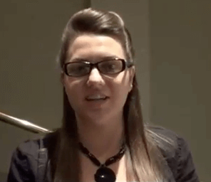 A woman with long brown hair and glasses smiles and waves at the camera in an indoor setting, welcoming plumbers, electricians, and HVAC technicians to the event.
