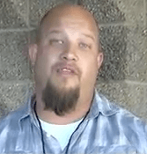 A man with a goatee and shaved head, dressed in a light plaid shirt, stands confidently in front of a textured block wall. He looks like an experienced electrician.