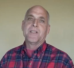 A middle-aged plumber in a red plaid shirt stands in front of a plain beige wall and looks at the camera.