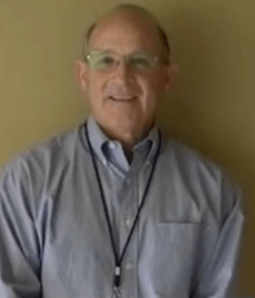 A man wearing glasses, a light blue button-down shirt, and a lanyard smiles at the camera as he stands in front of a plain beige wall. He looks ready to start his day as one of the local HVAC technicians.