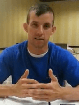 A man in a blue shirt—possibly a skilled service professional such as a plumber or electrician—sits at a table, clasps his hands, and speaks. A beige background is behind him.