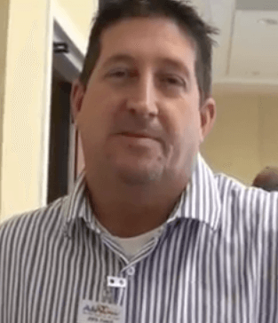 A man in a striped button-up shirt and a visible name tag stands indoors, facing the camera with a neutral expression. He projects the professionalism typical of HVAC technicians and service professionals.