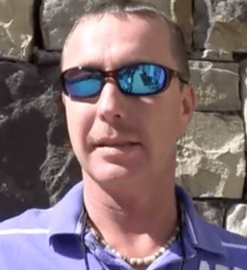 A man in reflective sunglasses and a purple collared shirt stands confidently in front of a stone wall, exuding the self-assured demeanor common among electricians and HVAC technicians.
