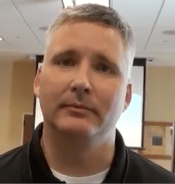 A man with short gray hair and a neutral expression stands indoors in front of a blurred background that shows a screen and ceiling lights. He looks like a typical plumber or HVAC technician.
