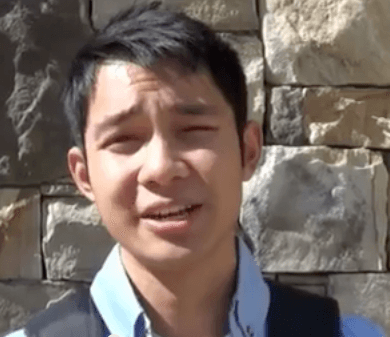 A young man with short dark hair—likely a local Service Professional—stands outdoors before a stone wall and squints slightly in the sunlight.