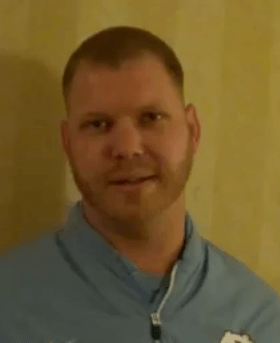 A man with short hair and a beard wears a light blue zip-up jacket and stands against a beige wall, looking like a skilled HVAC technician at work.