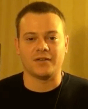 A man with short brown hair and a black shirt stands indoors in front of a yellow-beige background, representing skilled service professionals like electricians or plumbers.