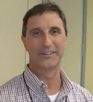 A man with short dark hair, dressed in a checked shirt and a lanyard, stands indoors and smiles at the camera, representing dedicated service professionals like electricians and plumbers.