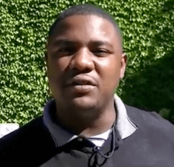 A man in a black collared shirt, similar to those plumbers or HVAC technicians wear, stands outside in front of green leafy plants.