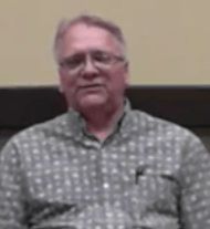 An experienced HVAC technician with glasses and short gray hair wears a patterned button-up shirt and sits in front of a beige and dark brown background.