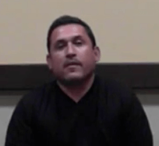 A man with short dark hair and a trimmed beard, dressed in a black shirt, sits against a beige and brown wall. He projects the professionalism typical of skilled service professionals such as plumbers or electricians.