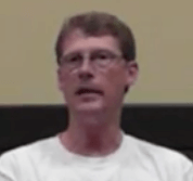 A man with short brown hair and glasses, wearing a white shirt that makes him look like an electrician, sits in front of a beige wall.