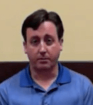 A man with short brown hair and a blue collared shirt sits in front of a beige and brown wall, looking straight ahead. He looks ready to assist as a trusted Service Professional.