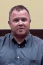 A man with short hair, dressed in a blue button-up shirt like many professional electricians wear, sits against a light yellow and brown background.