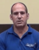 A man with short hair, possibly an electrician, sits against a beige and brown background and wears a blue collared shirt.