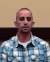 A man with a shaved head and trimmed beard, dressed in a blue and white plaid shirt, sits indoors against a tan and brown background. He looks ready to help as an expert service professional.