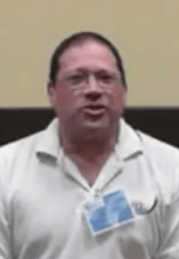 A man with glasses and short dark hair, dressed in a white collared shirt and blue name tag, sits in front of a beige and brown wall. He looks ready to assist you as one of the skilled plumbers on his team.
