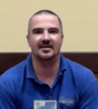 A man with short hair and a trimmed beard sits indoors against a beige and brown background. He wears a blue collared shirt, dressed like many professional plumbers.