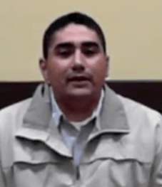 A man with short dark hair, wearing a light-colored jacket over a collared shirt, sits in front of a beige and brown wall. He looks like an experienced service professional—a reliable HVAC technician or trusted plumber.