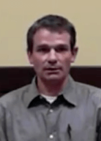 A man with short brown hair and a gray button-up shirt sits in front of a beige and dark brown background. He represents skilled service professionals such as electricians and HVAC technicians.