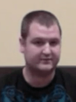 A man with short brown hair, light skin, and a neutral expression sits in front of a plain beige wall. He wears a dark shirt, and his attire and setting resemble those of many professional electricians.
