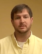 A man with short brown hair and a beard stands against a plain beige background, wearing a light yellow collared shirt that gives him the appearance of a professional plumber.
