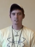 A young man stands in front of a plain beige background, dressed casually like many HVAC technicians on the job. He wears a light-colored T-shirt with a large graphic, a necklace, a lanyard, and a baseball cap.