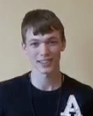 A young person with short brown hair stands in front of a plain beige background, wearing a black shirt with a white letter "A"—a style common among aspiring service professionals such as HVAC technicians and electricians.