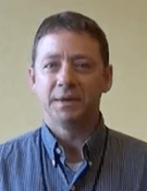 A man with short brown hair, dressed in a blue collared shirt typical of service professionals like plumbers, stands in front of a plain beige background.