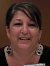 A dark-haired woman smiles confidently at the camera, wearing large skull-shaped earrings and a black top against a neutral brown background. Her bold style could inspire service professionals like plumbers or HVAC technicians to add flair to their own work attire.