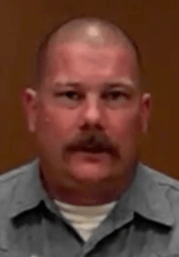 A man with a shaved head and mustache, wearing a gray collared shirt, sits in front of a brown background. He looks like an experienced HVAC technician.