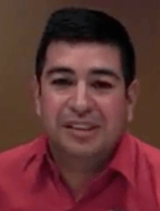 A man with short dark hair smiles at the camera. He wears a red collared shirt and stands against a brown background, representing Service Professionals such as skilled electricians.