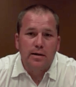 A man with short hair, dressed like an electrician in a white collared shirt, looks directly at the camera against a brown background.