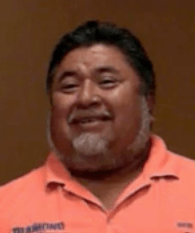 A man with short dark hair and a beard smiles as he wears a peach-colored collared shirt, giving him the professional appearance of many service workers, against a plain brown background.