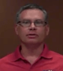 A man with short gray hair and glasses wears a red collared shirt and stands in front of a plain brown background, dressed like many HVAC technicians on the job.