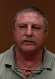 A middle-aged man with short gray hair and a mustache, who resembles many experienced HVAC technicians, stands against a plain brown background. He wears a gray collared shirt and has glasses hanging around his neck.