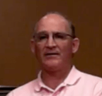 A middle-aged Service Professional with short hair and glasses wears a light pink collared shirt and stands in front of a plain brown background.