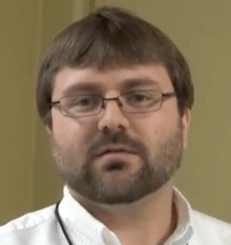 A man with short brown hair, glasses, and a beard wears a white collared shirt and looks directly at the camera against a plain indoor background, representing the professionalism of Service Professionals like electricians.