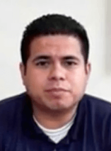 A man with short dark hair and a neutral expression faces the camera. He wears a dark shirt over a light undershirt, similar to the attire of electricians or other service professionals. The plain background is light.