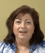 A woman with shoulder-length dark hair, dressed in a blue and white patterned top and a silver necklace, smiles warmly in front of a plain light green background—perfectly embodying friendly Service Professionals.