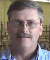 A middle-aged electrician with glasses, short gray hair, and a mustache stands indoors in front of industrial shelving.