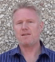 A man with short blond hair and a light beard wears a blue striped shirt and stands in front of a textured stone wall—typical attire for electricians or plumbers.