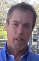 A middle-aged man with short graying hair, a goatee, and a blue collared shirt stands outdoors in front of trees and metal fencing. He looks like an experienced HVAC technician.