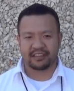 A man with short black hair, a trimmed beard and mustache stands outdoors in front of a stone wall. He wears a white collared shirt and a lanyard, projecting the confidence of an experienced electrician or service professional.