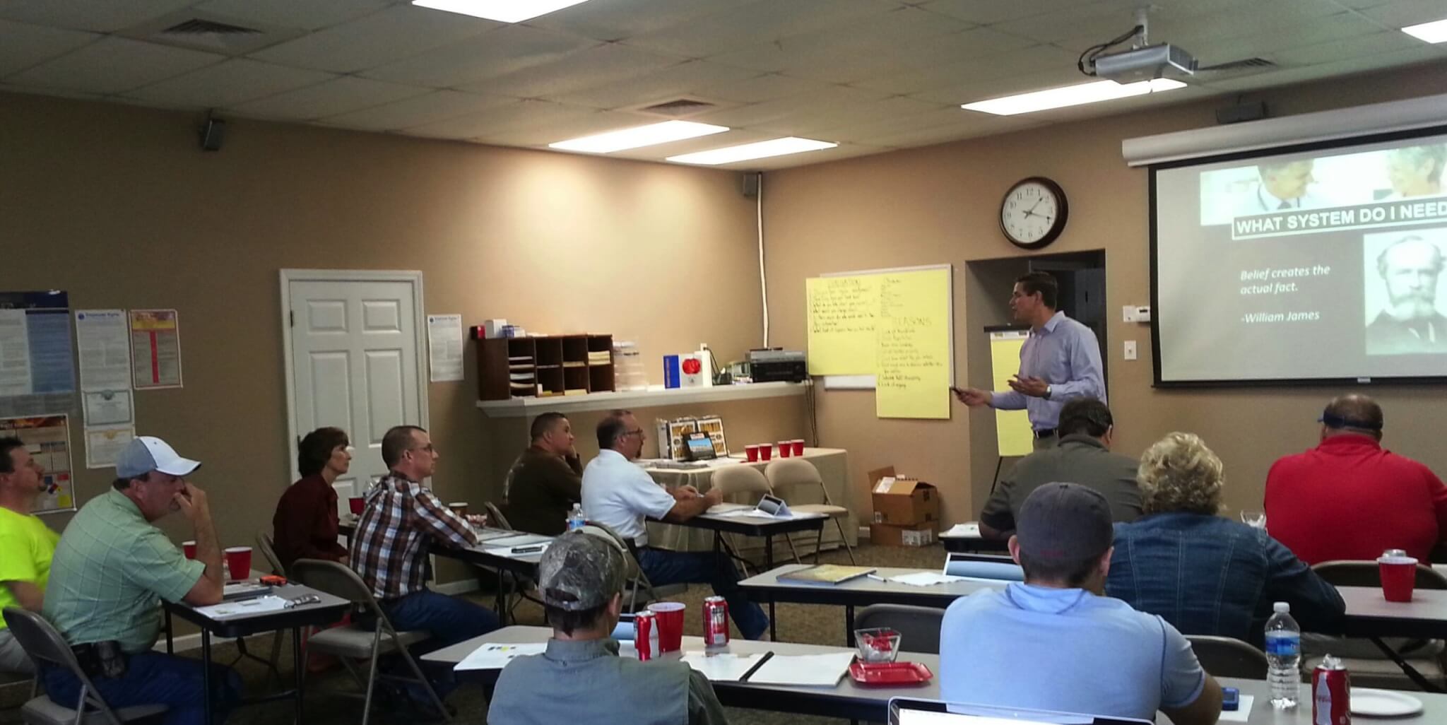 A man stands and presents to a group of HVAC technicians who sit at tables in a classroom. He projects his presentation on a screen at the front.