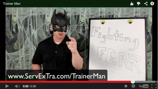A man wearing a Batman mask stands beside a whiteboard labeled "Fighting FEAR" and points directly at the camera—motivating service professionals such as plumbers and HVAC technicians. The website "ServExTra.com/TrainerMan" appears at the bottom.