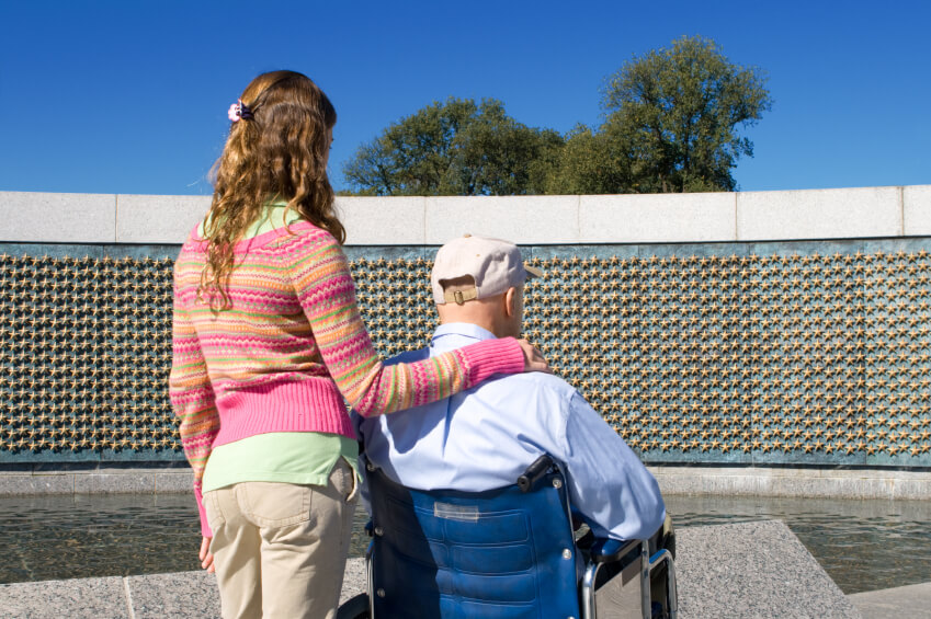 A young woman stands next to an elderly man in a wheelchair. They face a wall covered with gold stars at an outdoor memorial site that honors the dedication of service professionals like electricians and plumbers.