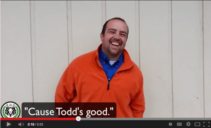 A man in an orange jacket smiles as he stands against a beige wall. The text on screen reads, 